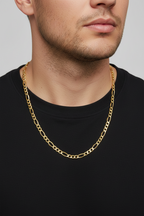 A close-up of a man wearing a gold Figaro chain necklace over a black t-shirt, against a neutral gray background.