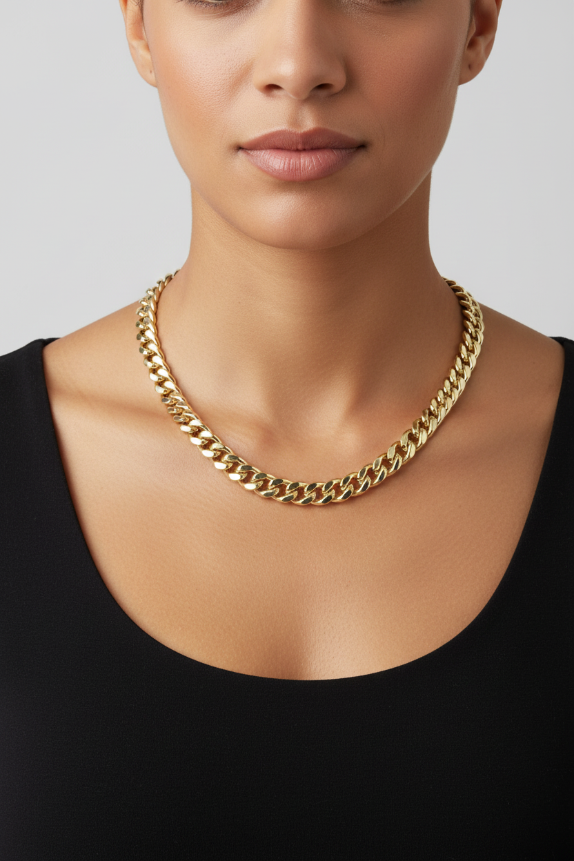 A close-up of a model wearing a thick, polished gold curb chain necklace with a black scoop-neck top against a light gray background.