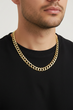A close-up of a man wearing a thick, shiny gold Cuban link chain necklace over a black crew-neck t-shirt, against a light gray background.