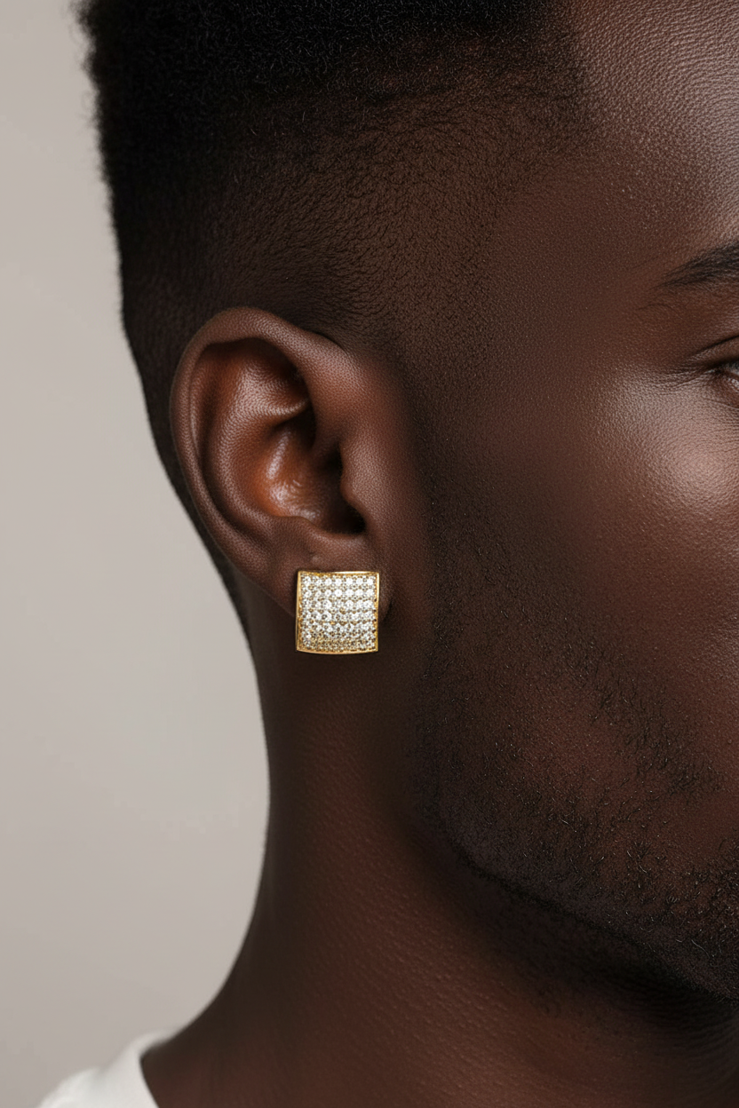 A close-up side profile of a man wearing a square gold earring paved with small clear stones. The earring is shown on his ear against a plain light gray background.