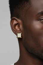 A close-up side profile of a man wearing a small square gold earring with a textured, diamond-like pattern on a light gray background.