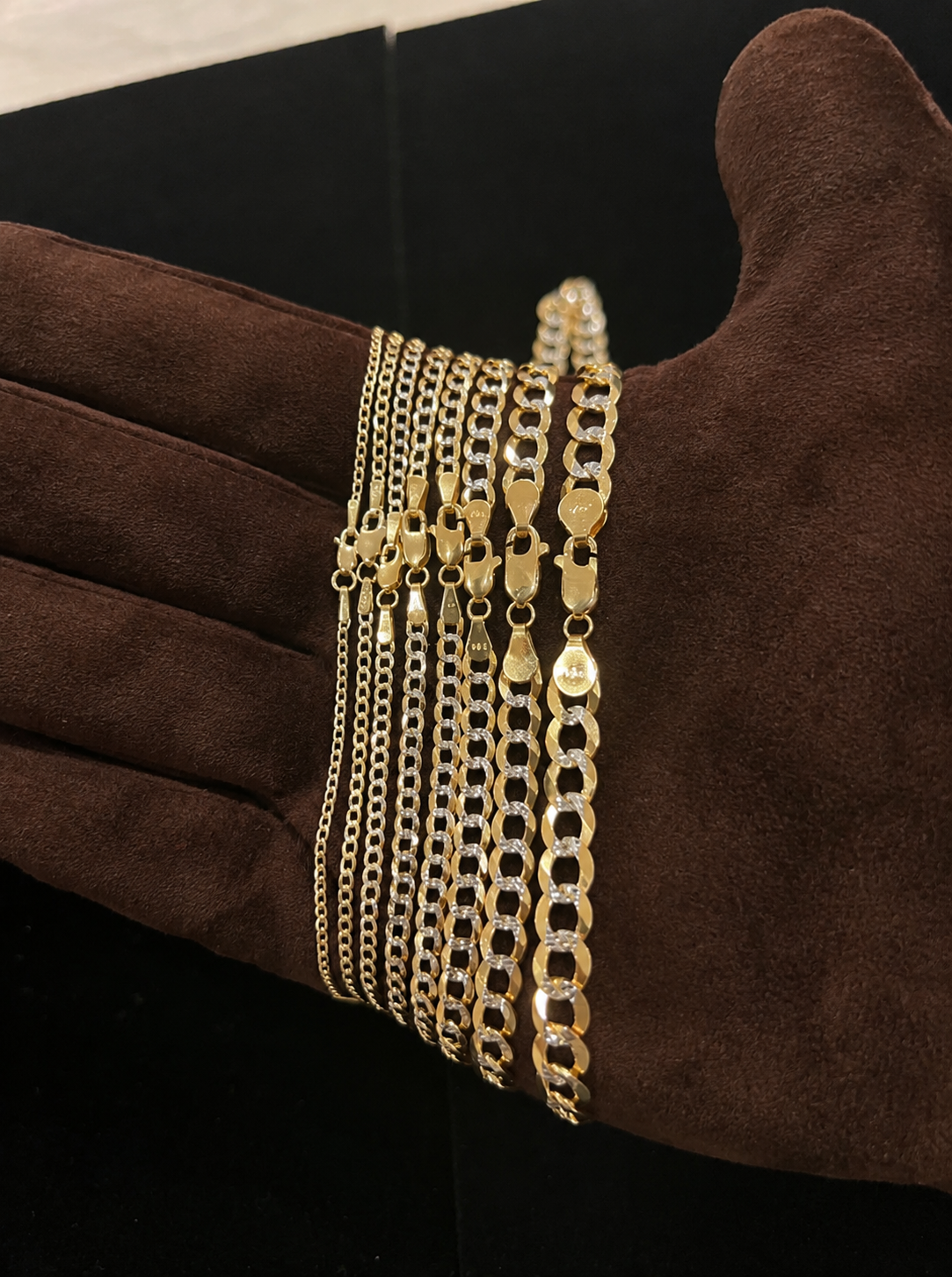 A close-up of several gold-tone chain necklaces displayed on a brown suede glove against a black background.